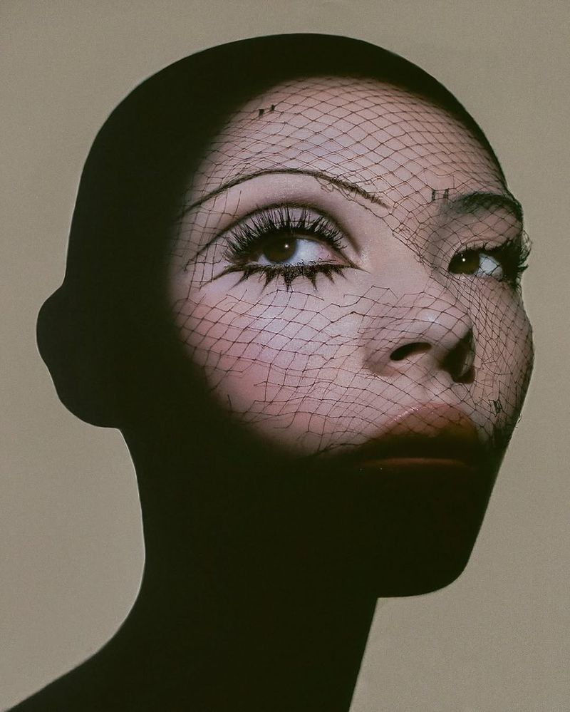 Glamorous portrait of an Asian woman with creative makeup and a black net veil, set against a neutral background.