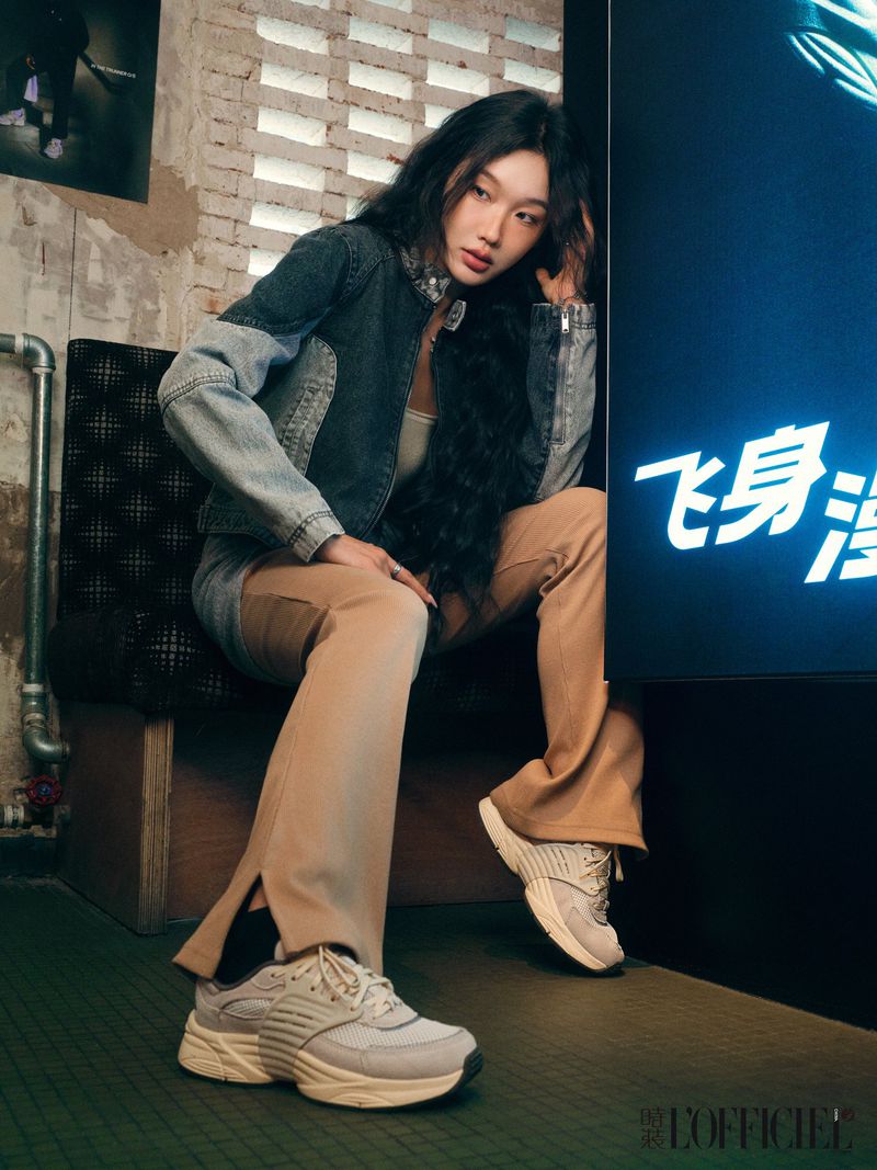 A stylish young woman sits in front of a bright sign in beige pants, a denim jacket, and sneakers, posing indoors with a brick wall.