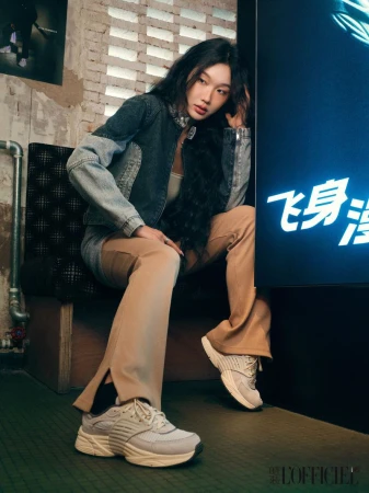 A stylish young woman sits in front of a bright sign in beige pants, a denim jacket, and sneakers, posing indoors with a brick wall.