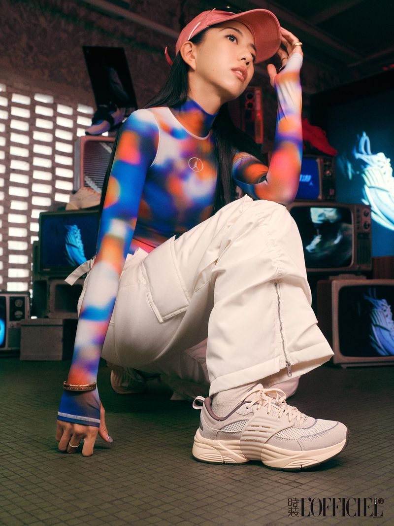 Asian woman in colorful top and white cargo pants poses with sneakers in a studio featuring vintage TVs and backlighting.