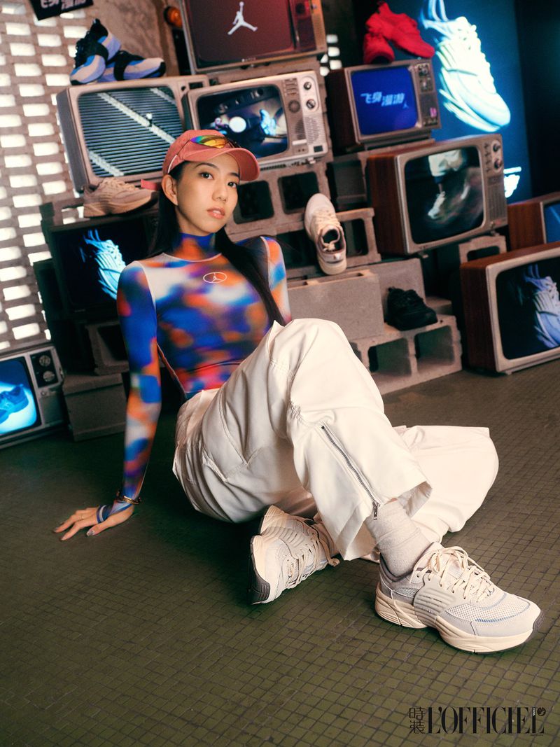 Stylish woman poses against a backdrop of stacked retro TVs wearing a tie-dye top, white pants, and sneakers in a studio setting.