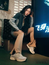 Woman in denim jacket and beige trousers seated against a textured brick wall with glowing signage