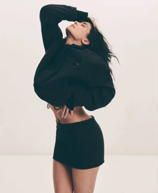 A woman with long dark hair poses with her head tilted back, wearing a black crop top sweatshirt and a black mini skirt in a studio shot.