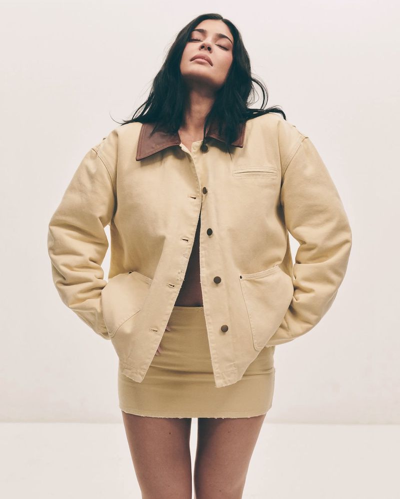 Woman in matching denim jacket and skirt poses with eyes closed against a white background in a fashion studio shot.