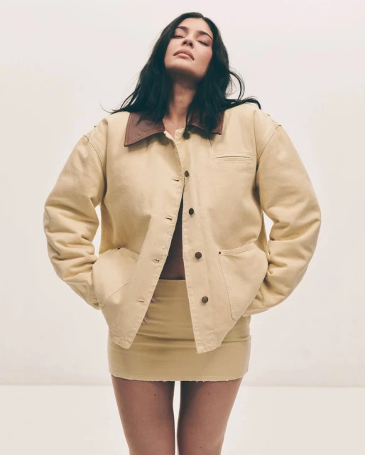 Woman in matching denim jacket and skirt poses with eyes closed against a white background in a fashion studio shot.