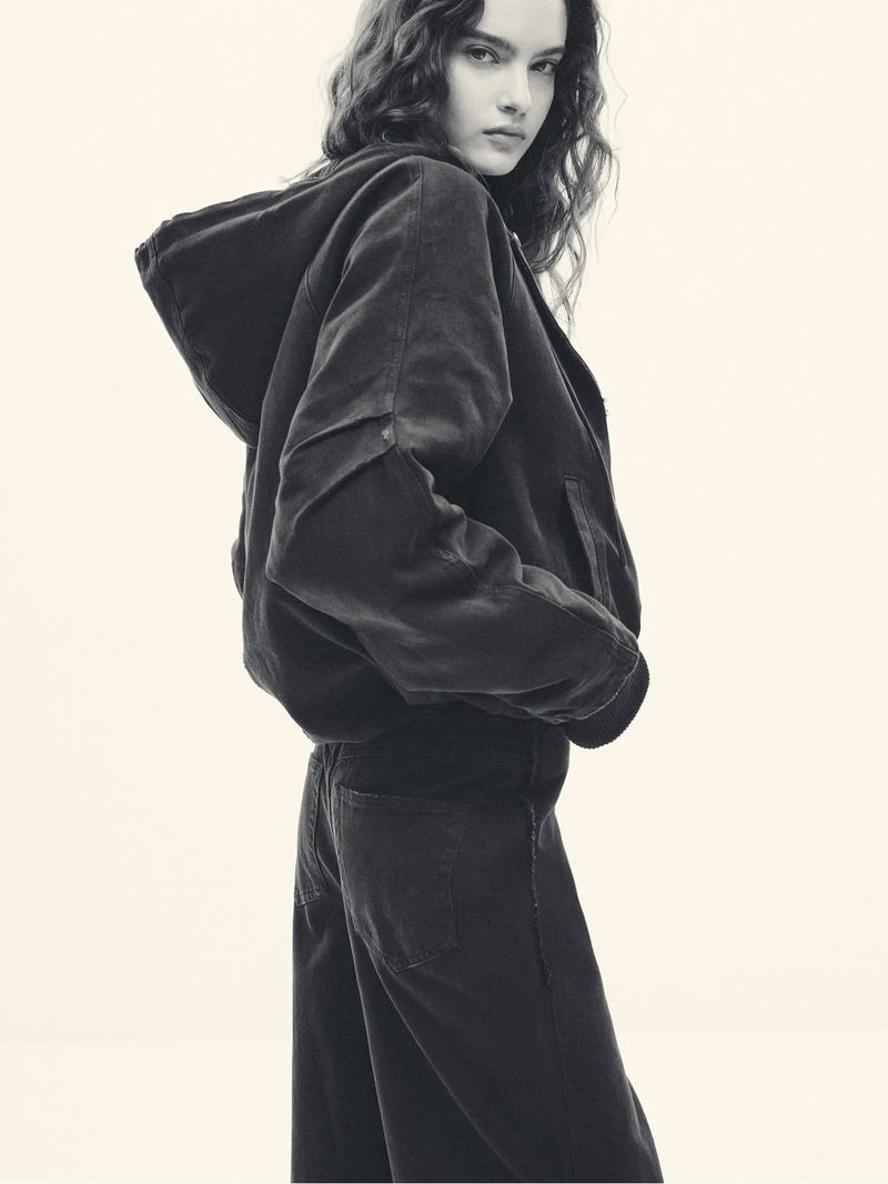 A young woman poses indoors, wearing a dark corduroy hooded jacket and matching jeans in a studio environment, with a neutral background.