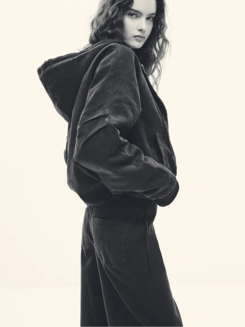 A young woman poses indoors, wearing a dark corduroy hooded jacket and matching jeans in a studio environment, with a neutral background.