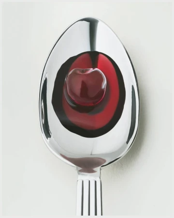 A single cherry sits in a pool of red liquid held in a shiny silver spoon, set against a plain white background in studio lighting.