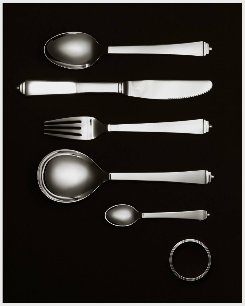 High-angle studio shot of symmetrically arranged silver cutlery: spoons, knife, fork, resting on a stark black background with a ring.
