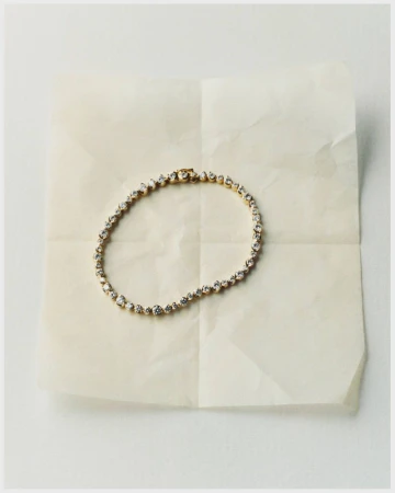 Elegant gold tennis bracelet with sparkling clear gems, displayed on a folded parchment paper background in a clean studio product shot.