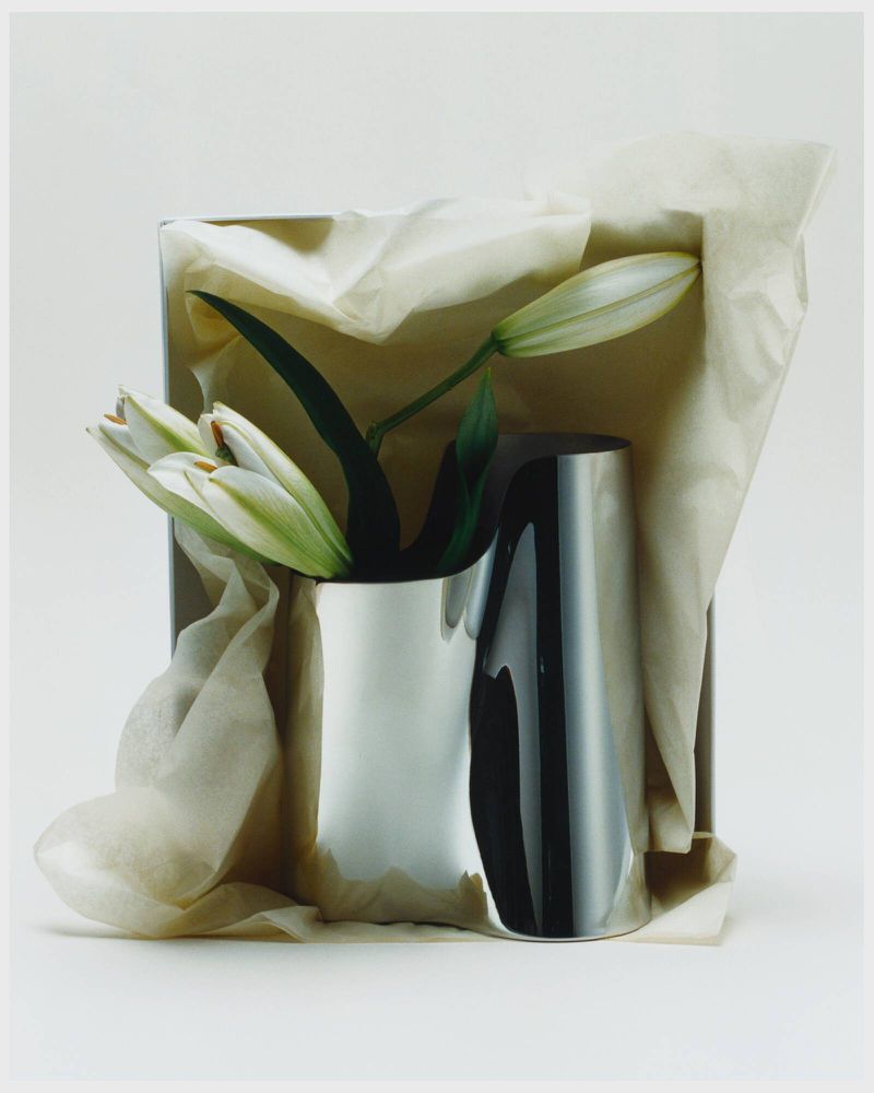 Still life shot of white lilies in a reflective silver vase, positioned in front of a gift box with folded tissue paper.