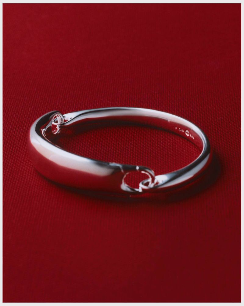Close-up of a silver-toned metal bangle bracelet with rounded edges against a deep red fabric background in a studio setting.