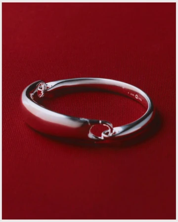 Close-up of a silver-toned metal bangle bracelet with rounded edges against a deep red fabric background in a studio setting.