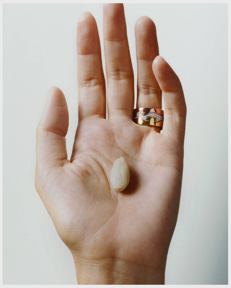 Close-up of a hand holding an almond against a white background. The hand features a ring on the ring finger.