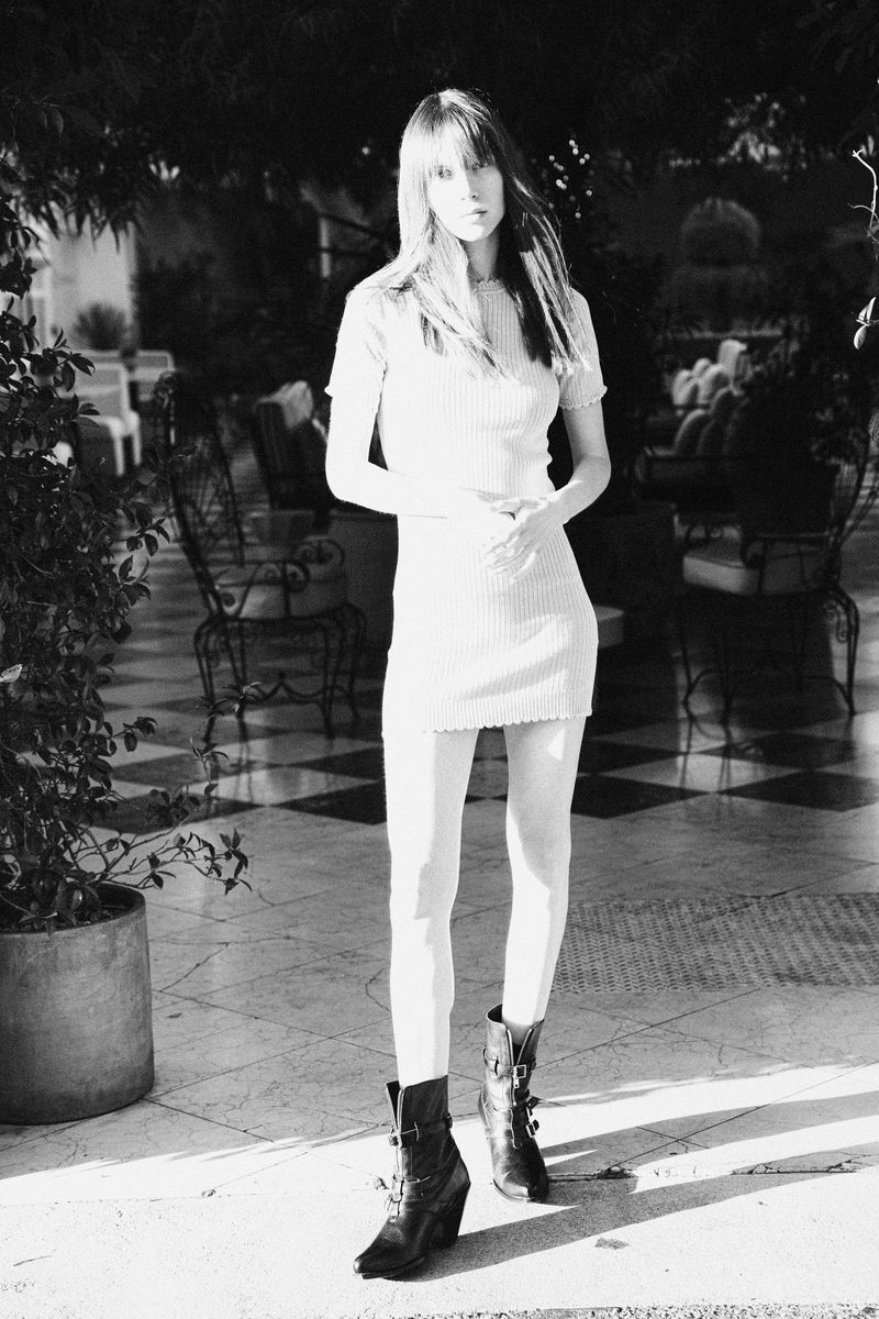 A long-haired young woman poses in a knitted dress and boots in a black-and-white fashion portrait taken outdoors on a checkered terrace.