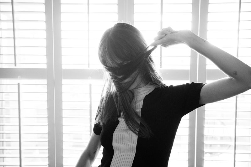 Monochrome portrait of a woman indoors pulling her hair over her face with her hand against a background of window blinds.