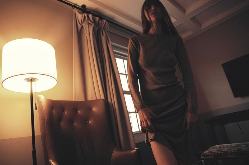 Woman in a dress poses indoors near a lamp and upholstered chair. Soft, warm lighting illuminates the scene, casting shadows on the wall.