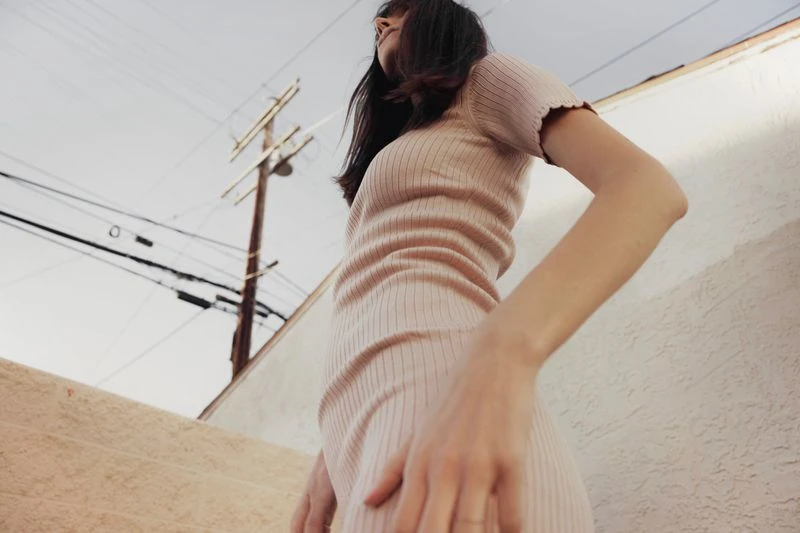 A woman in a pink dress stands outside near a white building and power lines, captured from a low angle on a partly cloudy day.