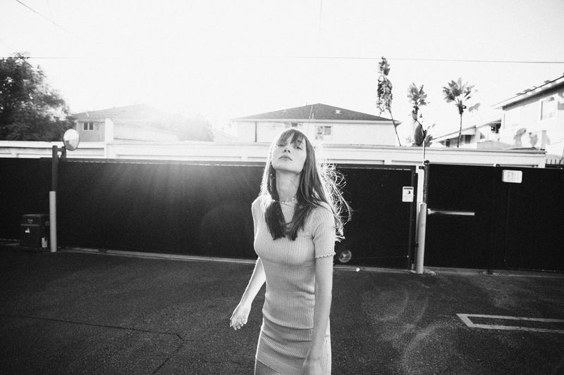 Black and white shot of a young woman with bangs in a short matching outfit in an urban setting with buildings in the background.