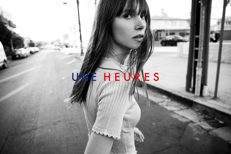 Woman with long brown hair is captured in a medium shot on a street, with cars parked and a building in the background.