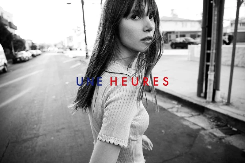 Woman with long brown hair is captured in a medium shot on a street, with cars parked and a building in the background.