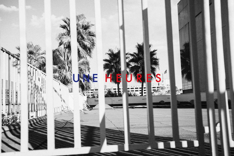 Black and white image features silhouetted palm trees seen through a white metal fence. Text 'UNE HEURES' overlayed in blue and red.