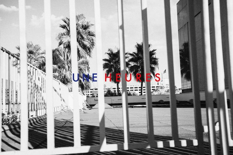 Black and white image features silhouetted palm trees seen through a white metal fence. Text 'UNE HEURES' overlayed in blue and red.