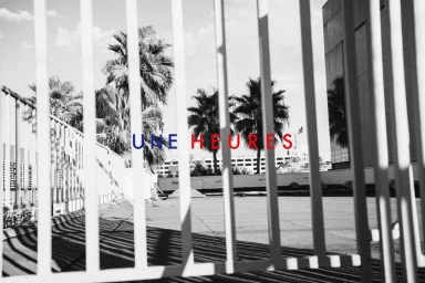 Vertical metal fence bars casting shadows across an outdoor rooftop terrace with palm trees and typography overlay reading UNE HEURES.
