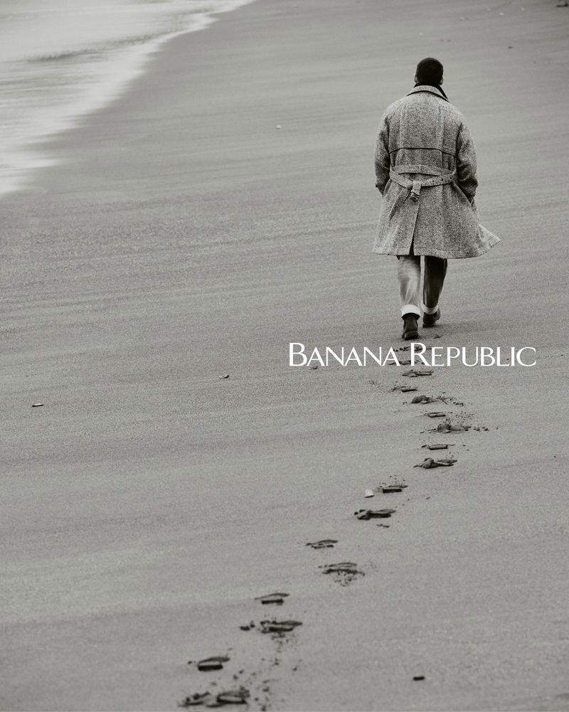 A man in a coat walks along a sandy beach, leaving footprints; fashion advertisement for Banana Republic in black and white.