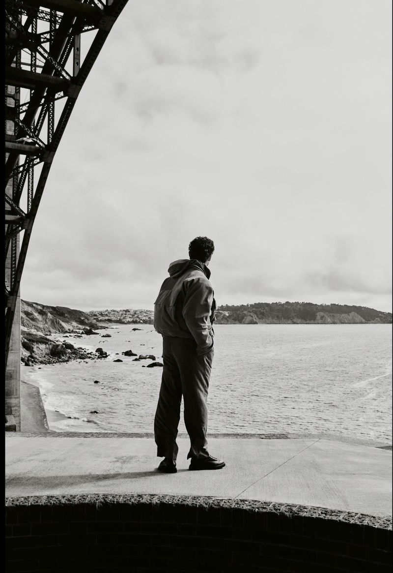 A person stands on a concrete platform by the sea, gazing out at the ocean, with a bridge structure visible in the background.
