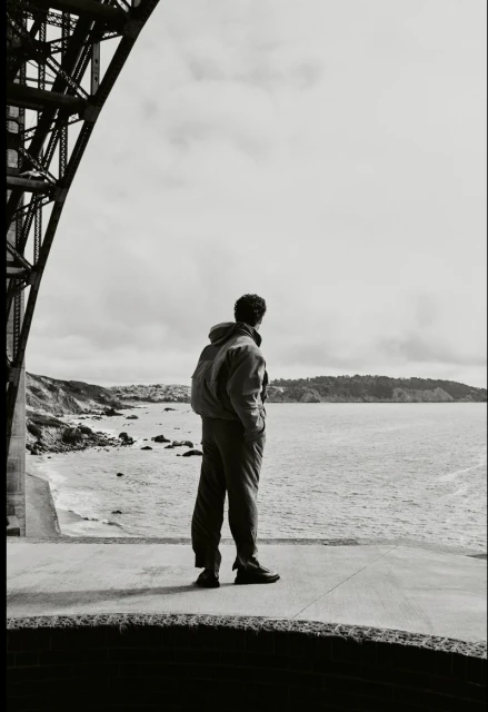 A person stands on a concrete platform by the sea, gazing out at the ocean, with a bridge structure visible in the background.