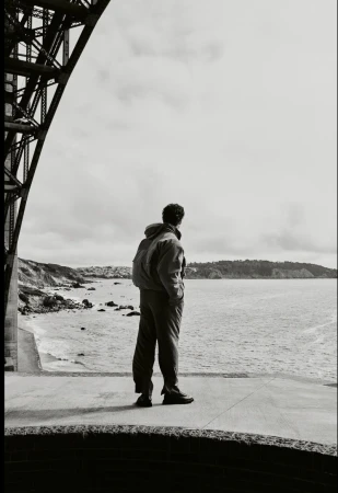 A person stands on a concrete platform by the sea, gazing out at the ocean, with a bridge structure visible in the background.