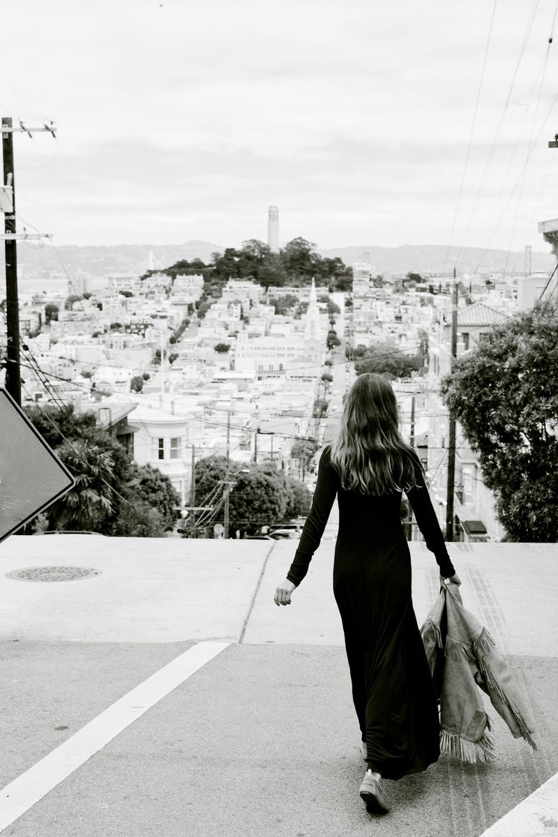 A woman in a long dress walks away on a city street in a black and white, eye-level shot with a cityscape in the background.