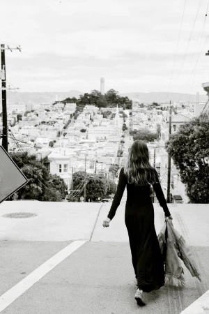 A woman in a long dress walks away on a city street in a black and white, eye-level shot with a cityscape in the background.