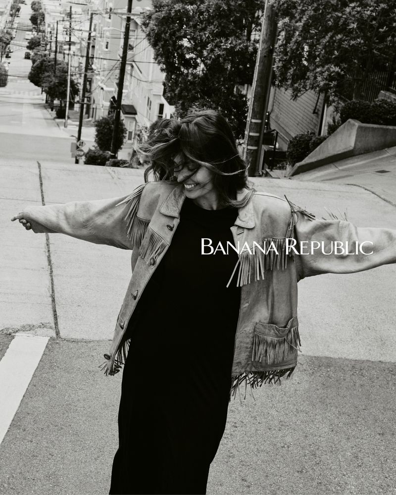 Woman wearing a fringe jacket dances on an angled street in a black and white outdoor shot; Banana Republic logo in the left center.