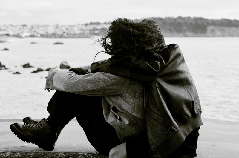 Black and white photo of two people embracing near the water wearing a wool coat, leather jacket, and lace-up boots.
