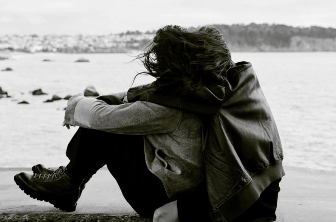 Black and white photo of two people embracing near the water wearing a wool coat, leather jacket, and lace-up boots.