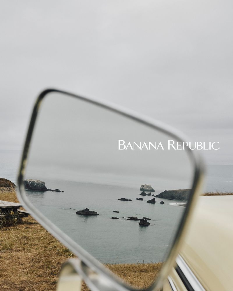 Scenic ocean view with rock formations reflected in a vintage car mirror under an overcast sky, featuring Banana Republic branding.