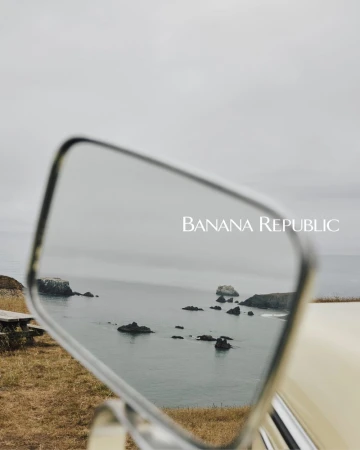 Scenic ocean view with rock formations reflected in a vintage car mirror under an overcast sky, featuring Banana Republic branding.