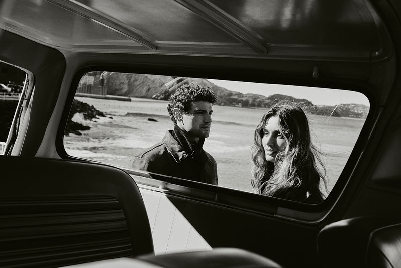Monochromatic shot from inside car facing couple near ocean. Man in jacket looks left, woman with wavy hair glances right near rocky coast.