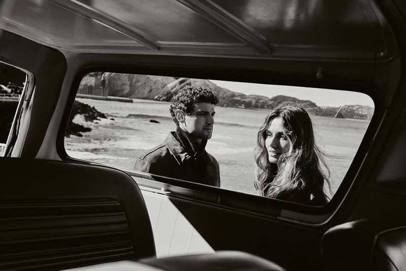 Monochromatic shot from inside car facing couple near ocean. Man in jacket looks left, woman with wavy hair glances right near rocky coast.