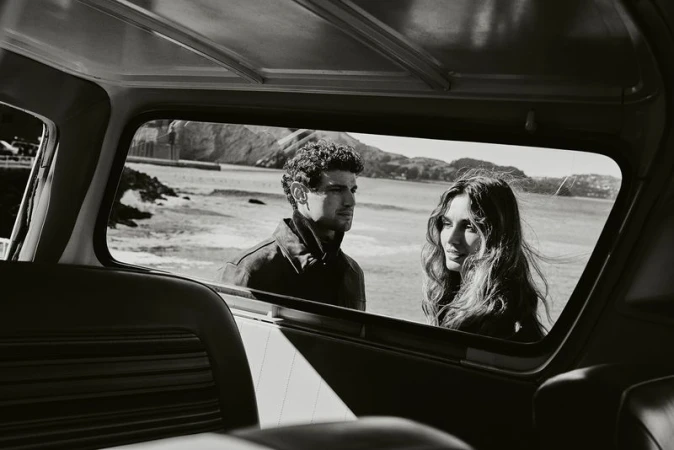 Monochromatic shot from inside car facing couple near ocean. Man in jacket looks left, woman with wavy hair glances right near rocky coast.