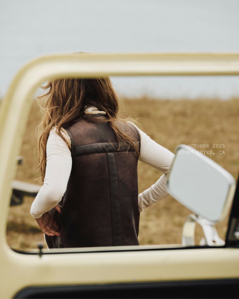 Woman in vest standing near vehicle in field, seen through vehicle window. Tan field and cloudy sky in background.