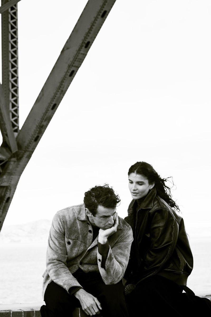 Monochrome shot of a seated man and woman in leather jacket on a bridge with open sky, looking towards each other in a pensive moment.