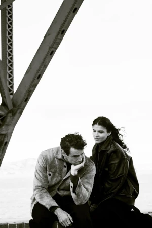 Monochrome shot of a seated man and woman in leather jacket on a bridge with open sky, looking towards each other in a pensive moment.