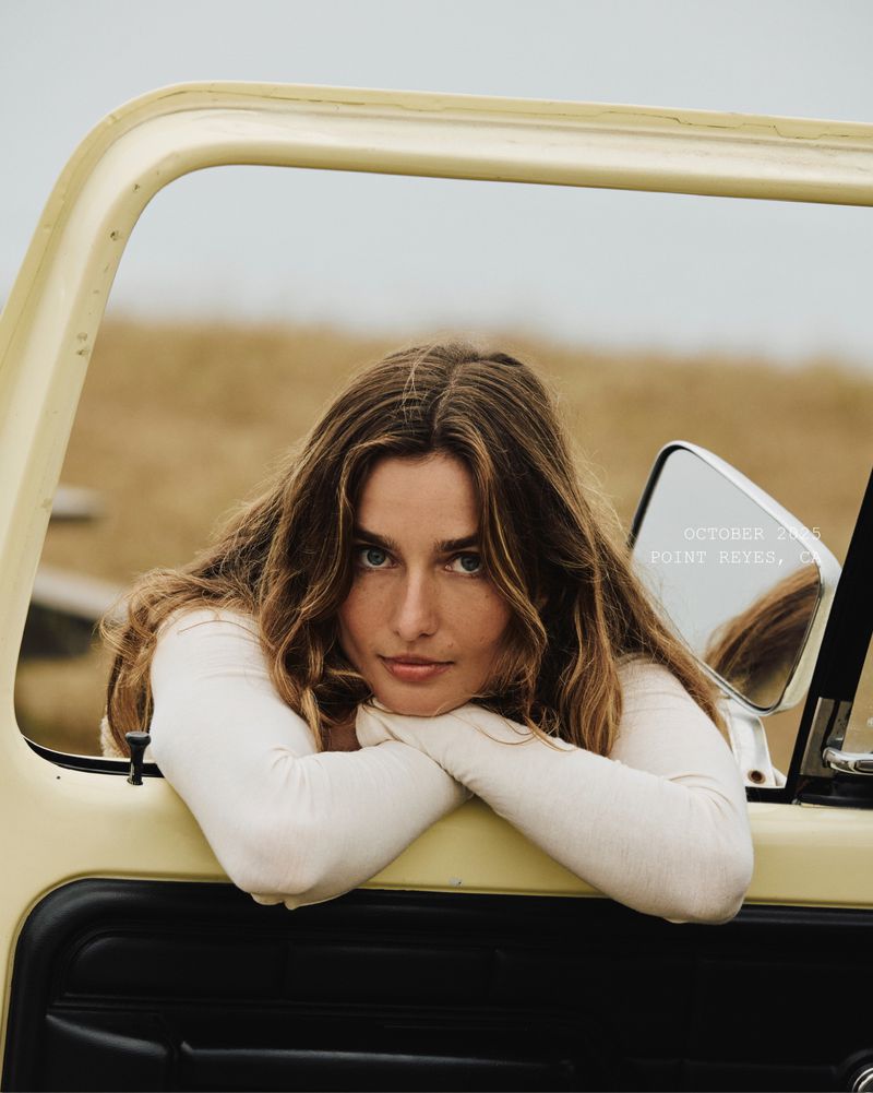 Young woman with long brown hair rests her arms on a car door, looking directly at the camera against a muted landscape backdrop.