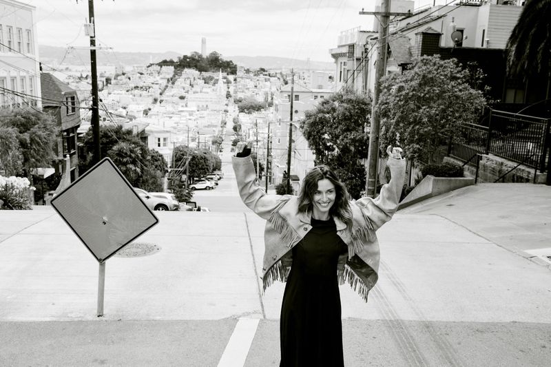 Woman in fringe jacket celebrates on San Francisco street with scenic cityscape, creating an exciting lifestyle fashion editorial.