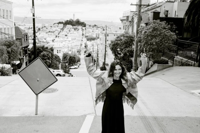 Woman in fringe jacket celebrates on San Francisco street with scenic cityscape, creating an exciting lifestyle fashion editorial.