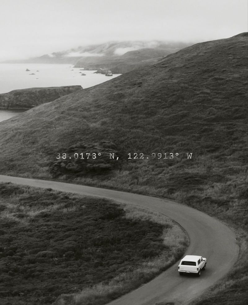Grayscale landscape featuring a winding road, a car, rolling hills, and the ocean under an overcast sky with geographical coordinates.