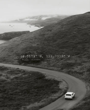 Grayscale landscape featuring a winding road, a car, rolling hills, and the ocean under an overcast sky with geographical coordinates.